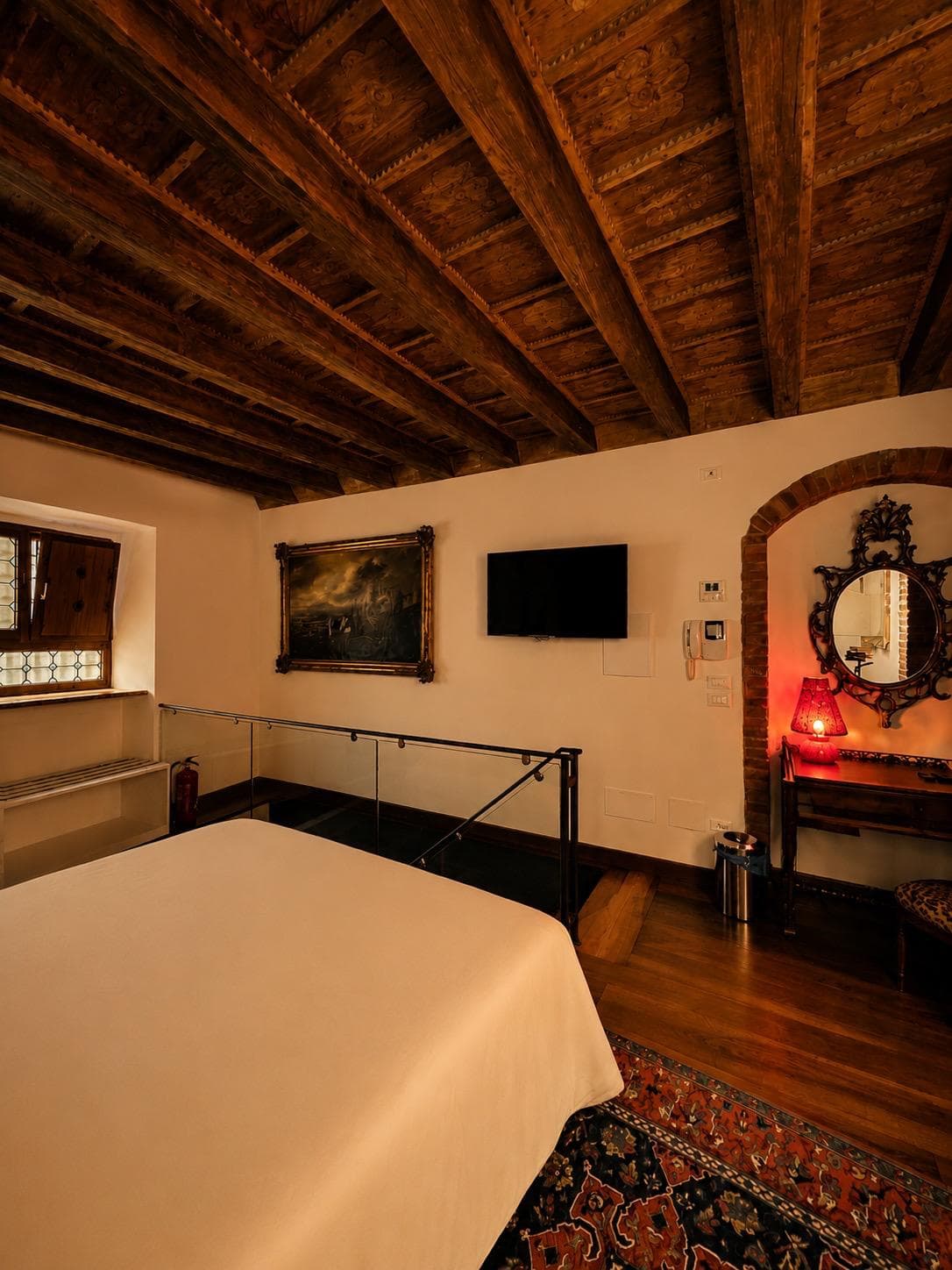 Soppalco della suite Marcello a Verona: tela paesaggistica del Settecento in cornice dorata accanto alla Smart TV con accesso Netflix gratuito, balaustra in vetro e soffitto a travi a vista