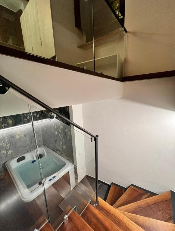 Scala in legno con balaustra in vetro che dalla suite Marcello al piano terra scende alla SPA privata al piano −1, vista dall'alto, con la vasca idromassaggio in basso e carta da parati botanica